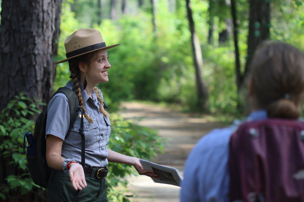 A smiling park ranger gesturing toward a hiker on a trail in the forest.