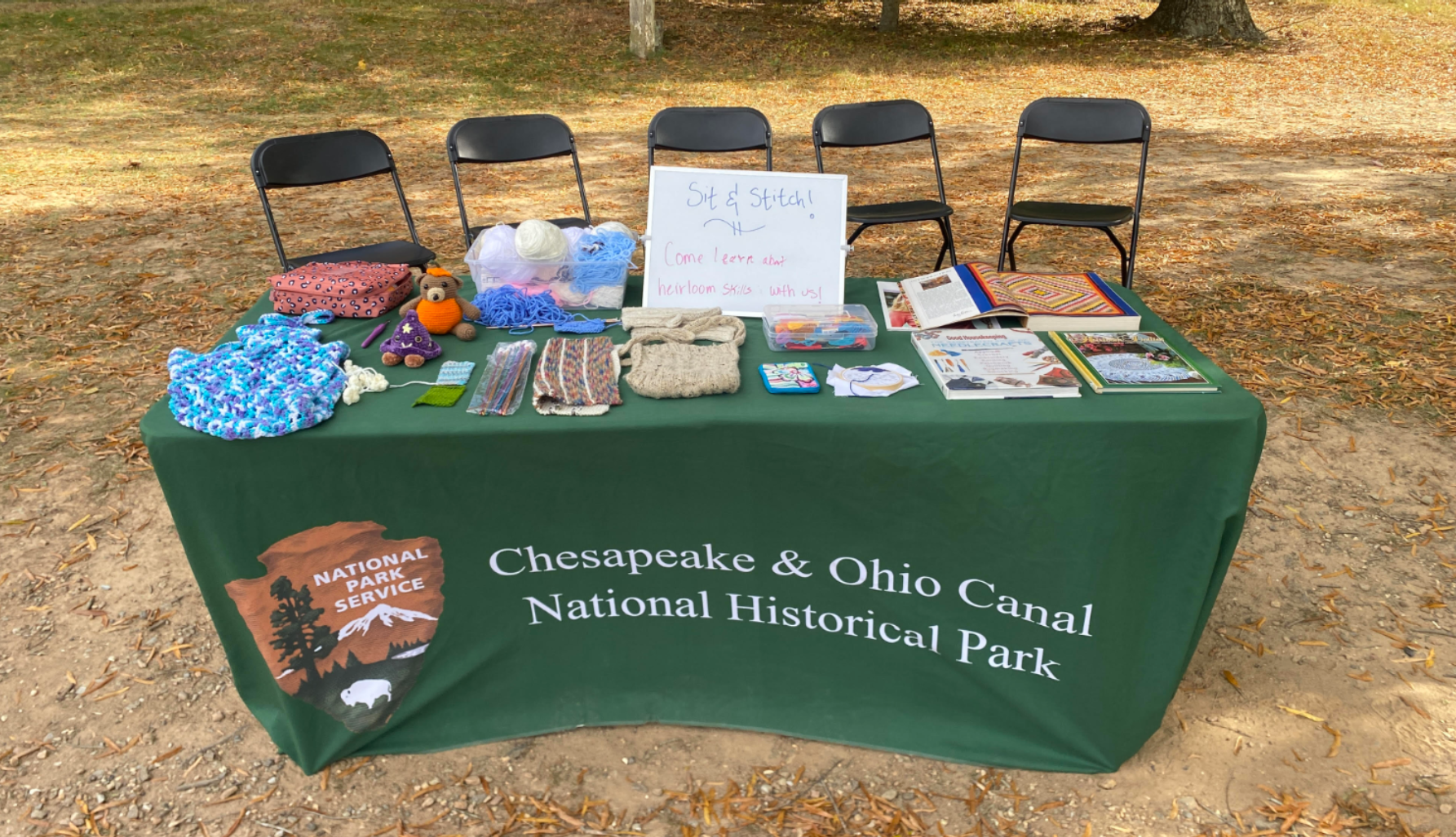 Table with a green cloth covered in needlework supplies, knitted scarves, and knitted animals