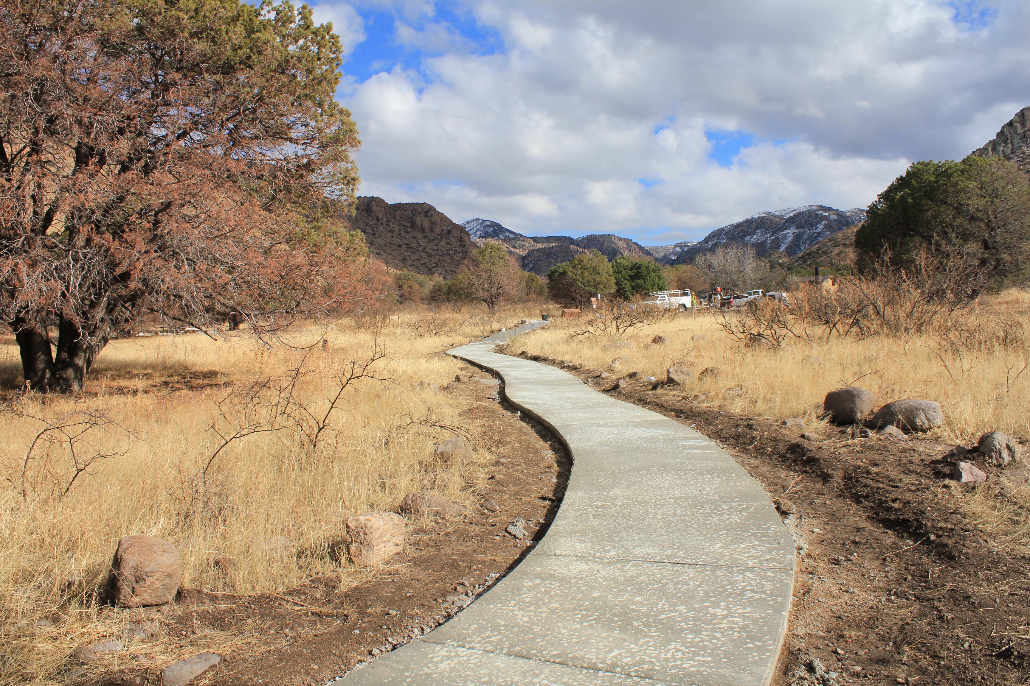 A winding concrete path curves through a dry grassland surrounded by scattered trees and rock.