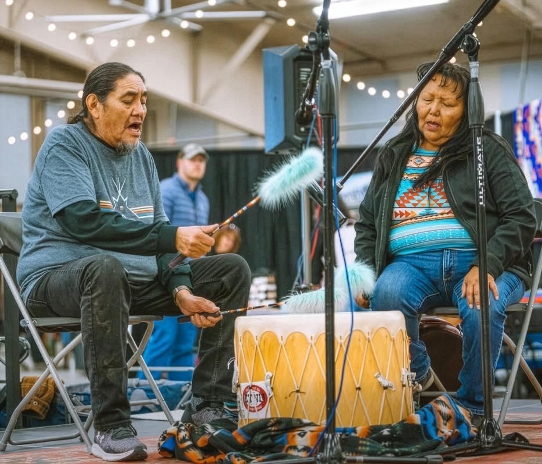 A man and a woman are seated. He is playing a large drum. Both are singing.