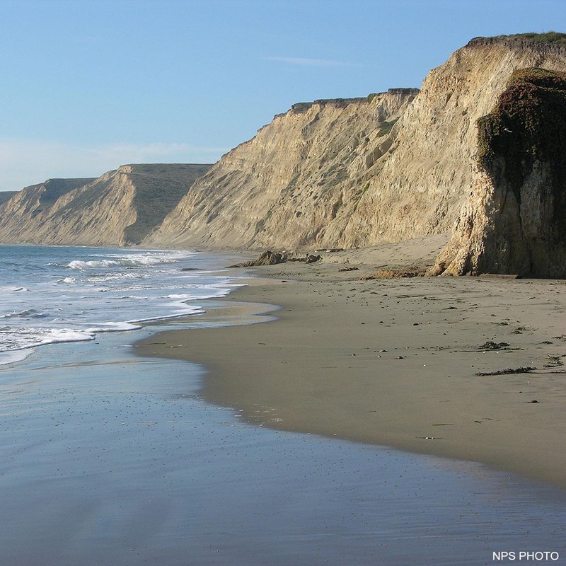 Cream-colored bluffs rise on the right above a sandy beach as waves wash in from the left.