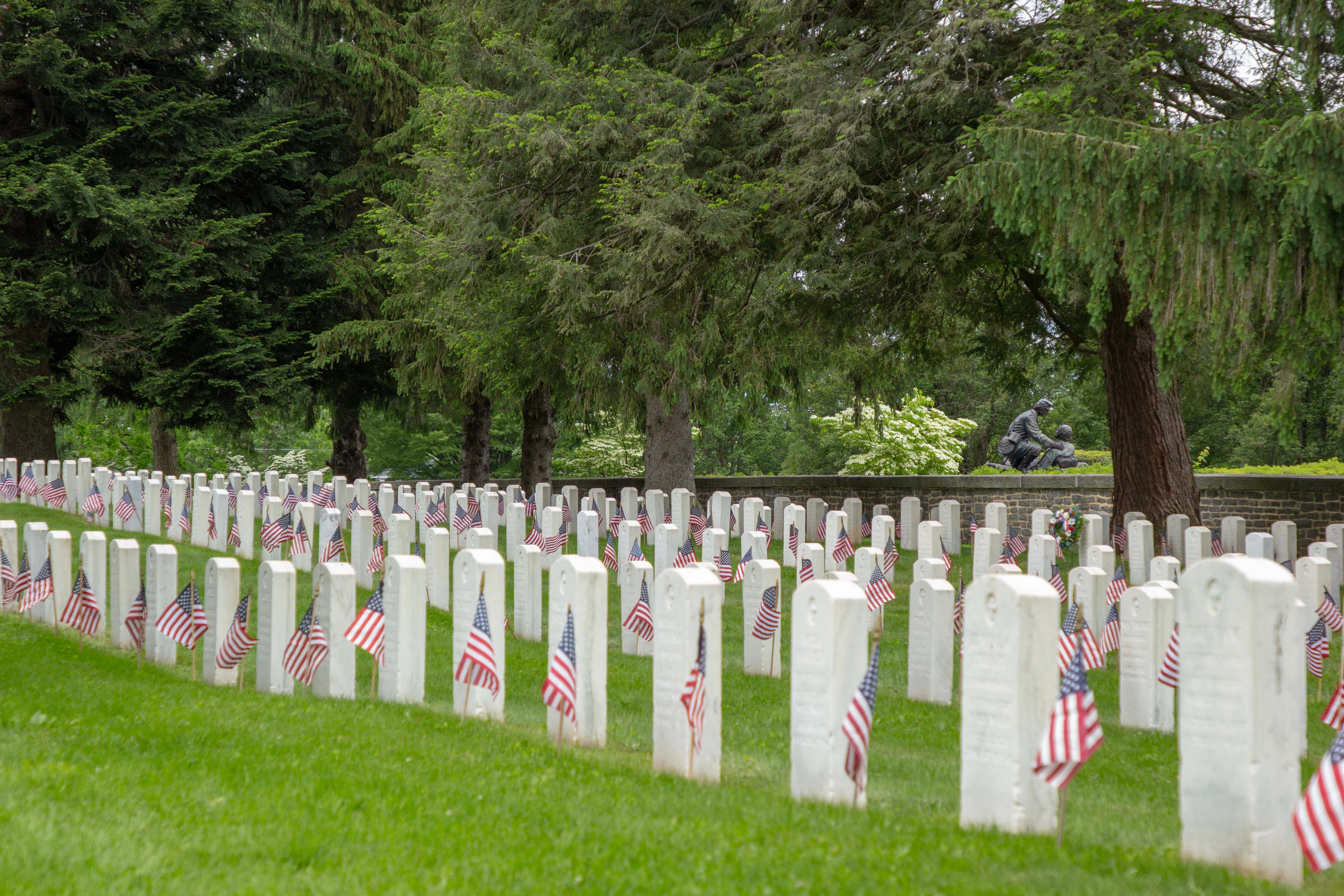 A cemetery with vertical white grave stones are decorated with small American flags.