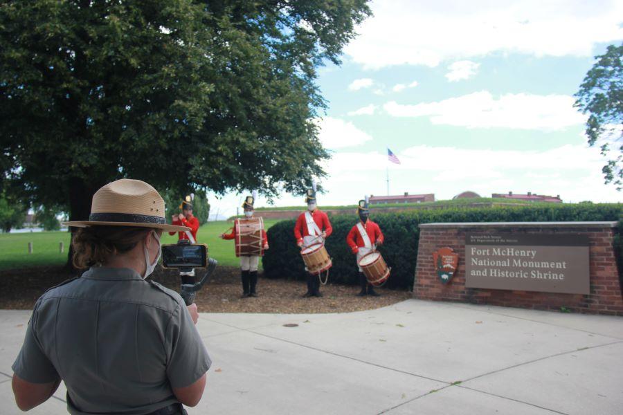 A park ranger films the Fort McHenry Fife and Drum Corps next to the Fort McHenry park sign.