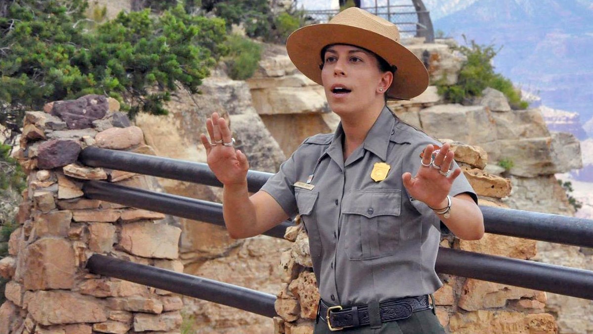 A park ranger in uniform is presenting a geology talk at a scenic overlook with guardrails.