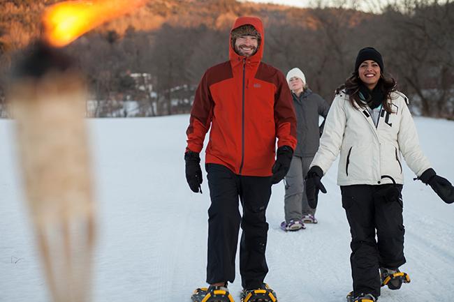 people in winter clothing walk towards camera wearing snowshoes with torchlight in foreground