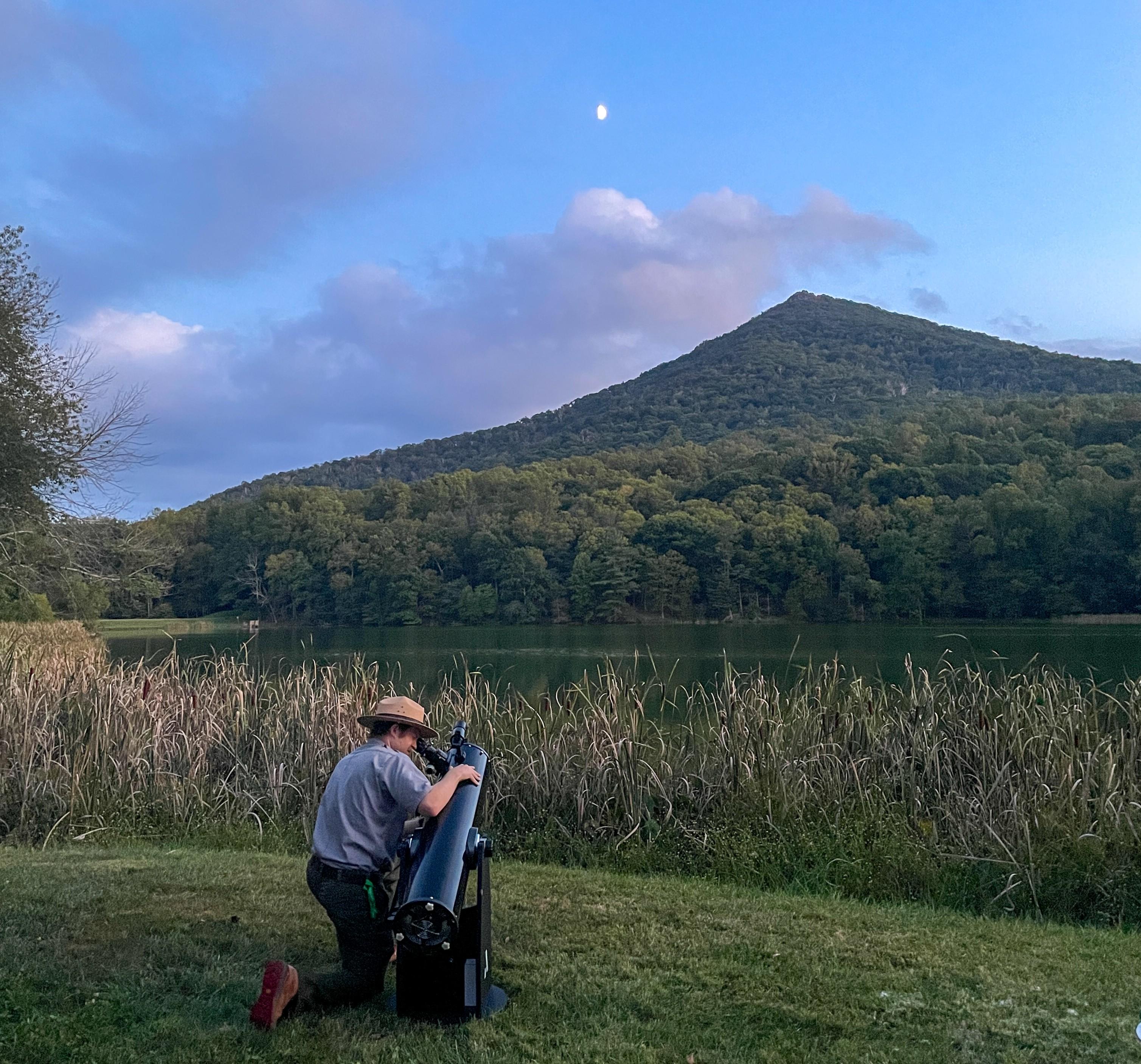 uniformed ranger kneels at a telescope pointing at the moon with a mountain in the background