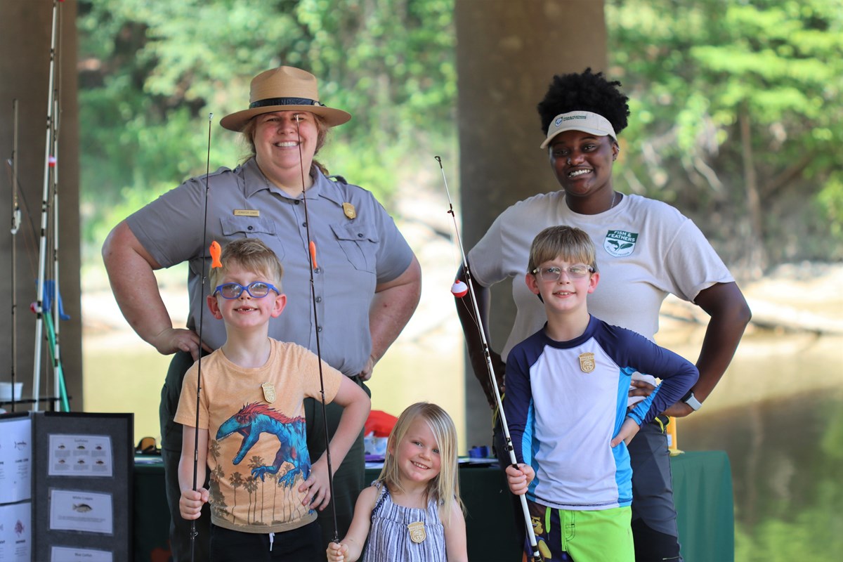 3 kids stand in front of 2 park rangers and hold fishing poles.