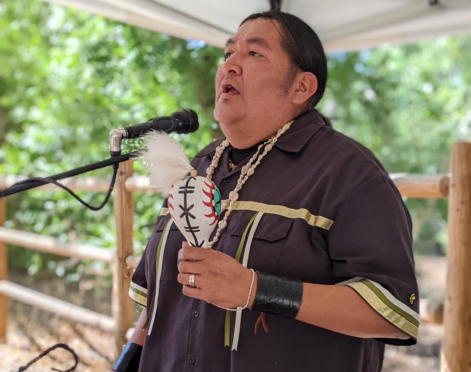 A middle-aged Hopi man is holding a rattle and singing into a microphone