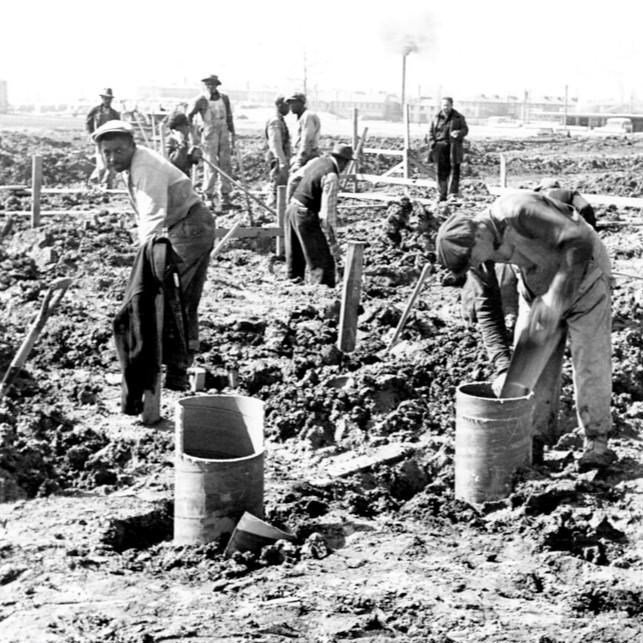 A group of Black and White construction workers stand in the mud building a foundation.