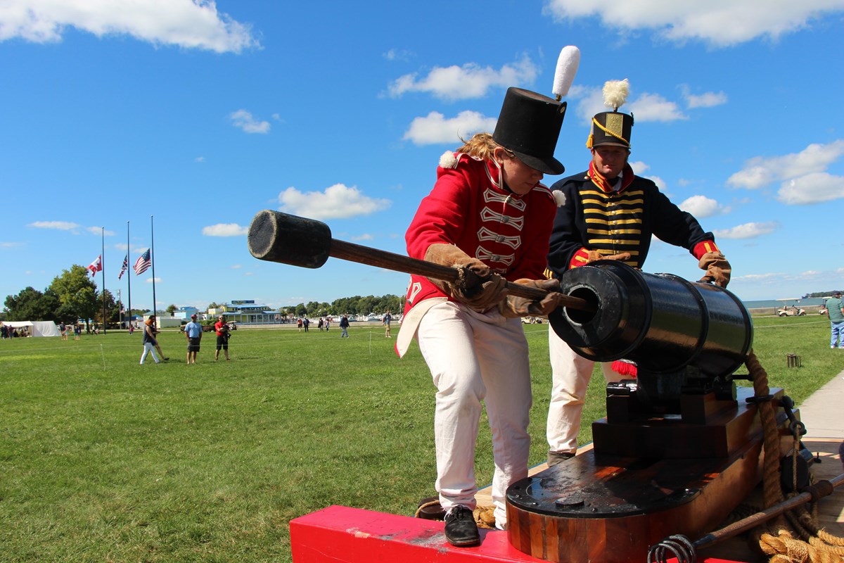2 people in historic uniforms stand next to cannon. One at front holds sponges. Other covers vent.