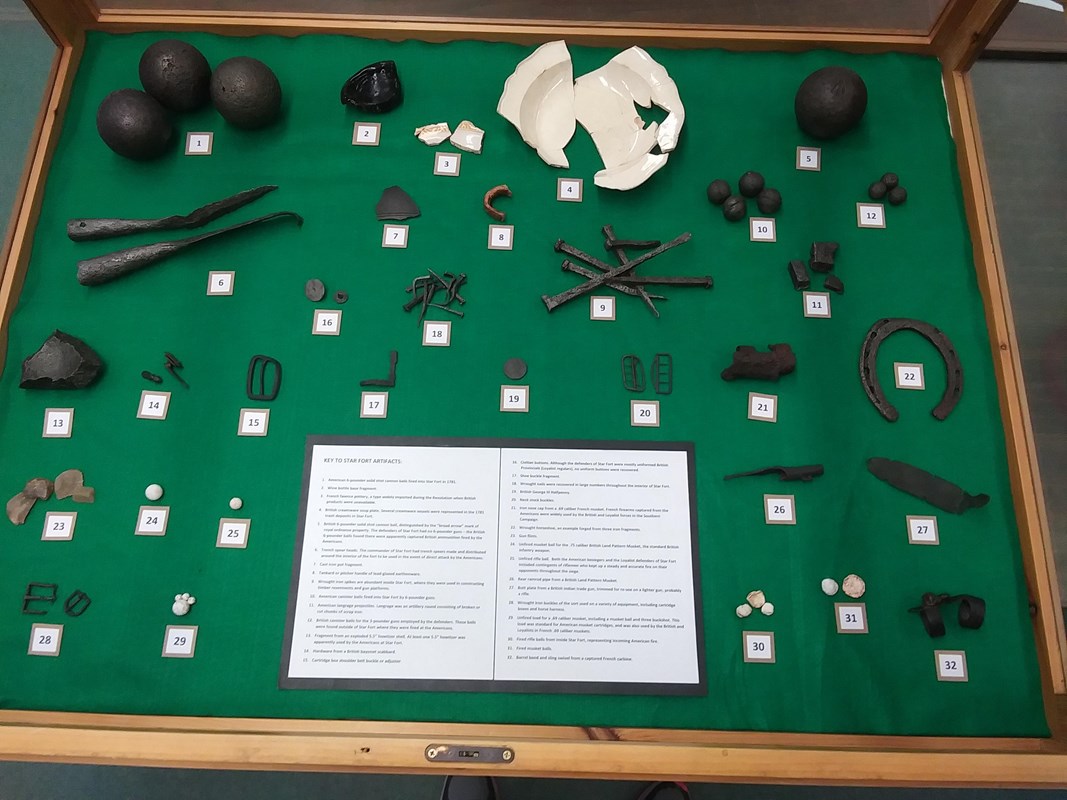 A collection of artifacts such as musket balls, nails, a horseshoe, a broken plate and cannon balls.