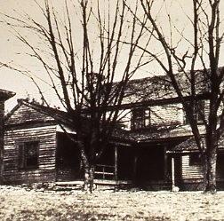 A black and white photo of a house, with several bare trees in front of it