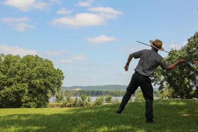 Image of the back of a park ranger throwing a spear with an atlatl on a green lawn.