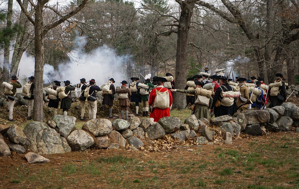A group of militia soldiers fire their muskets from behind a stone wall