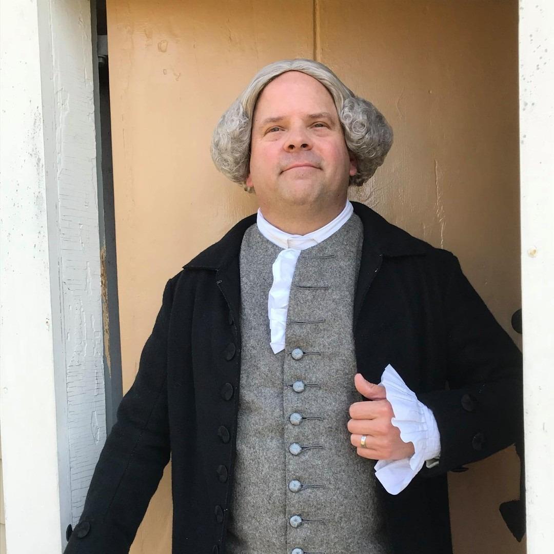 A man wearing a suit and wig in the style of the 1700s stands in the doorway of a historic home