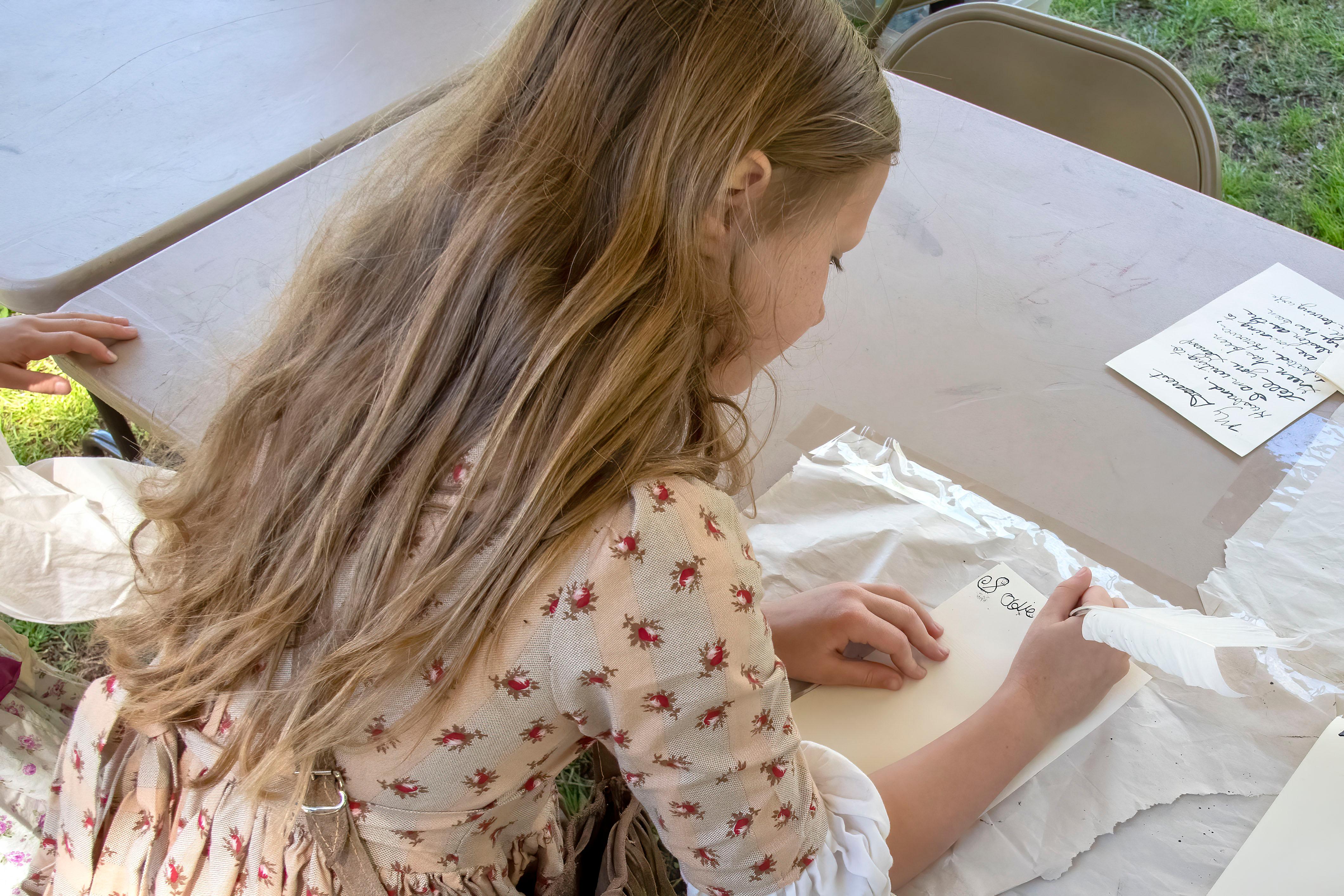 A young girl leans over a table as she writes with a feather quill.