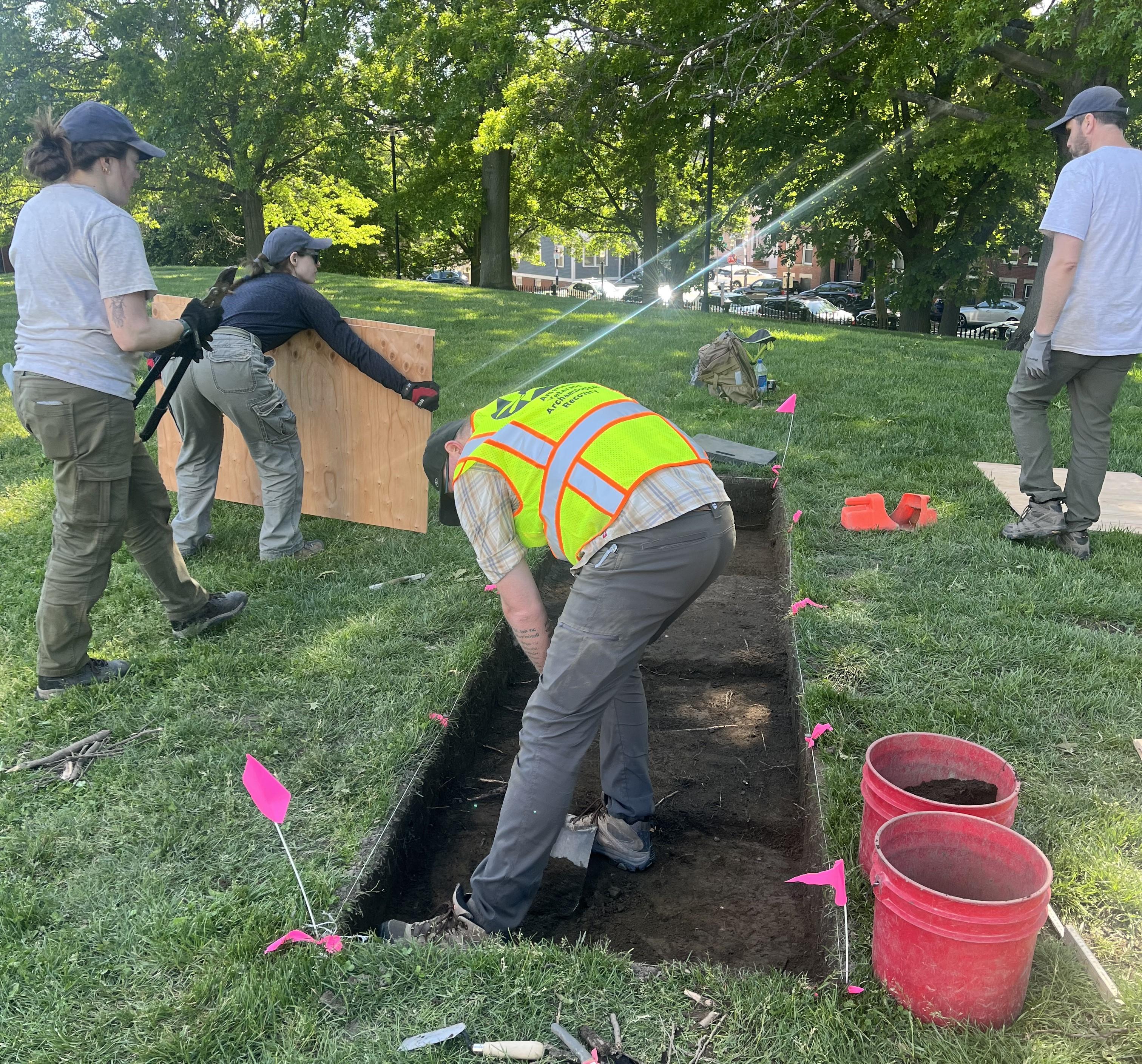 A person in a yellow vest stands in rectangular dug-up dirt area in a grassy lawn.