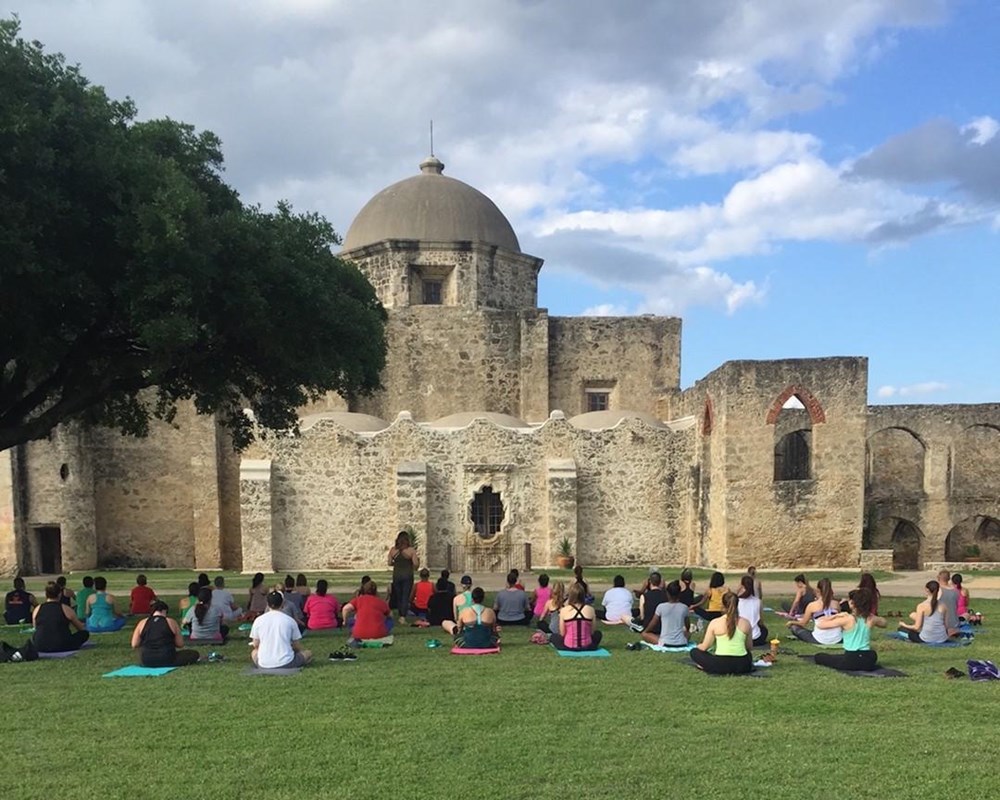 People sitting on the grass practicing yoga in front of large mission church.