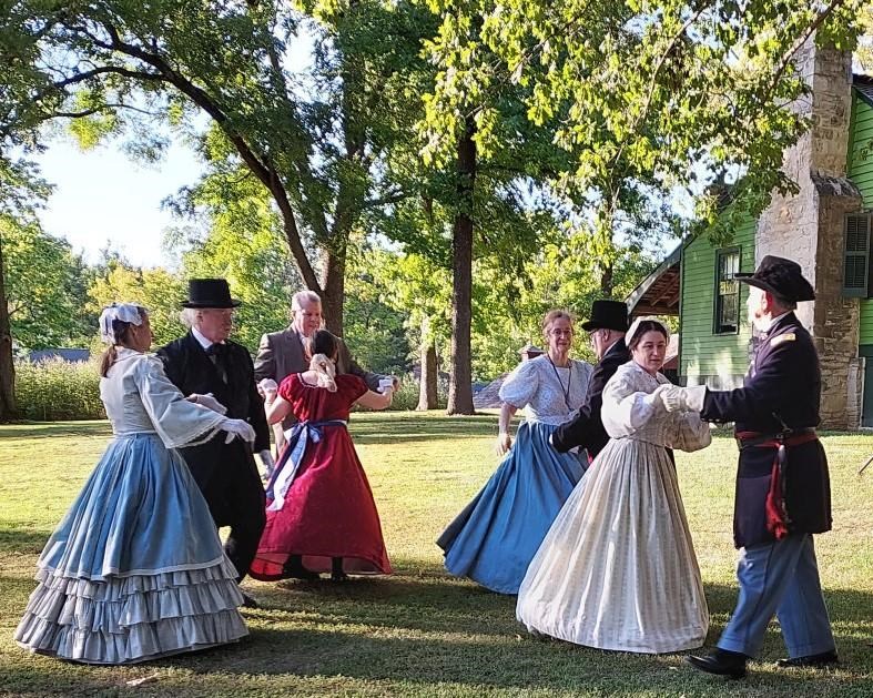 six people wearing Civil War era clothing dancing outside with trees and green house behind them