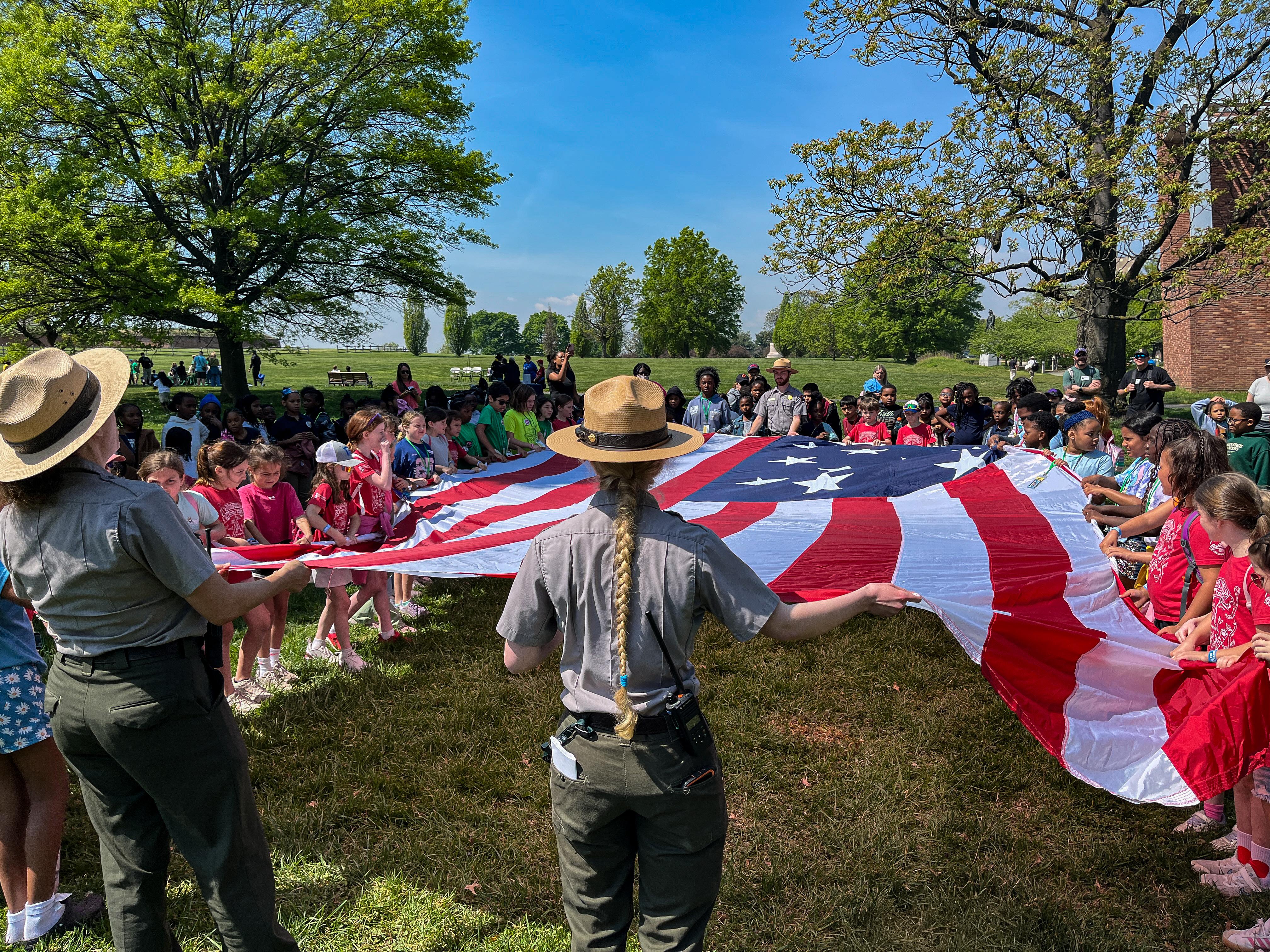 Two park rangers and a group of visitors hold the edges of a large American flag.