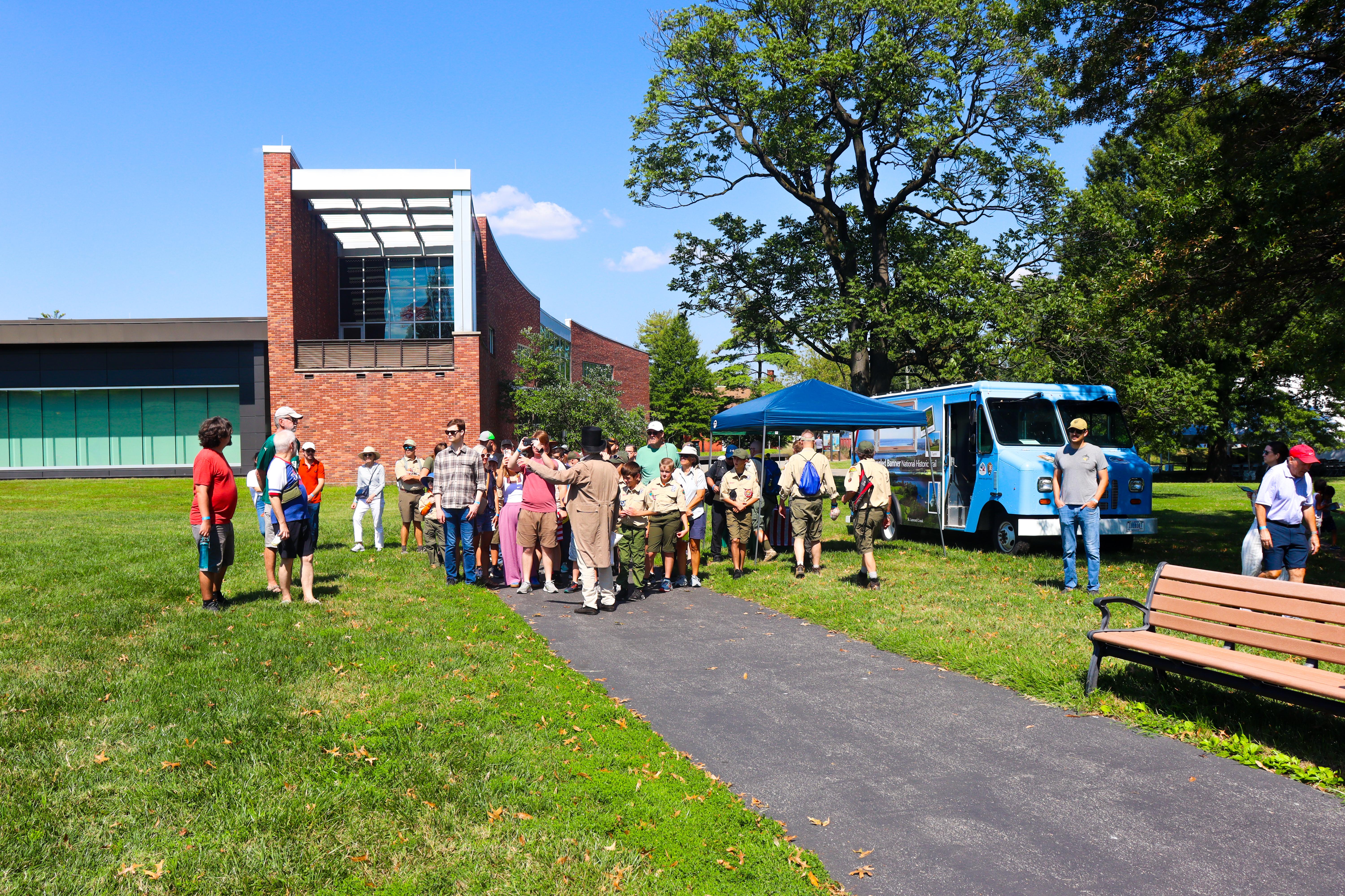 Ranger dressed in 1812-era garb leads a group of visitors up a paved path from a brick building.