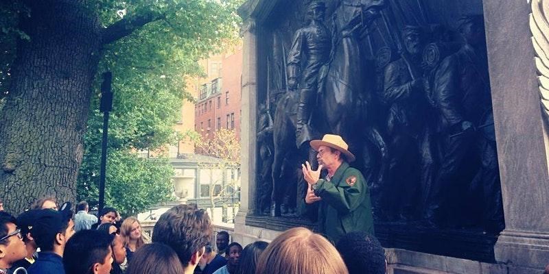 An NPS Ranger speaking to a group of children in front of a monument, holding an image of soldiers.