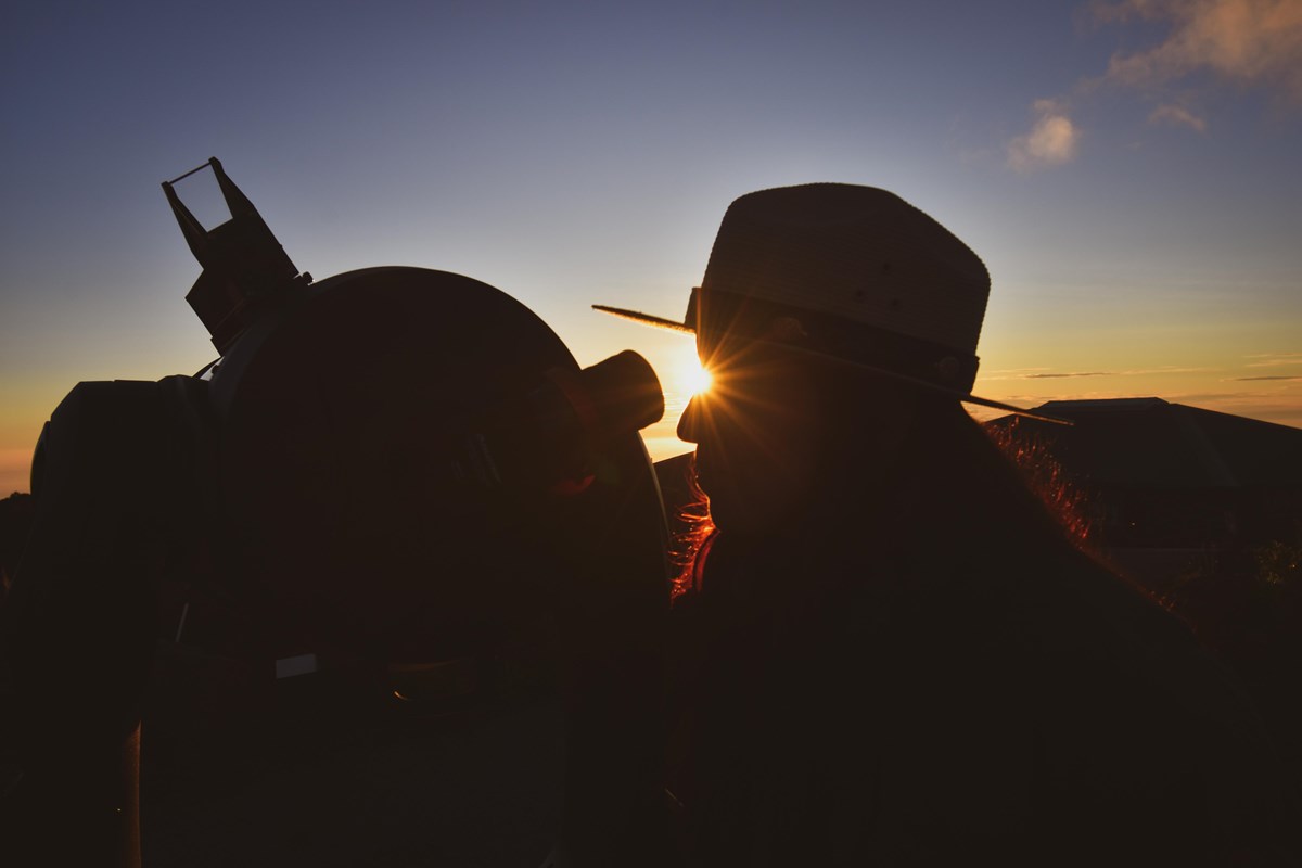 A ranger looks into a telescope with the sunsetting in the background.