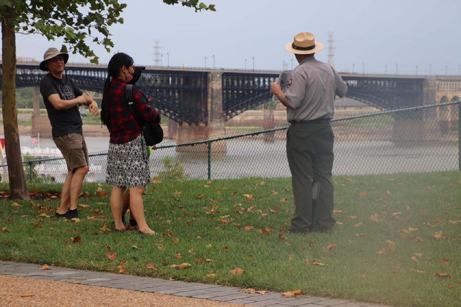 A man in a National Park Service ranger uniform gestures at a bridge. Two people look on.