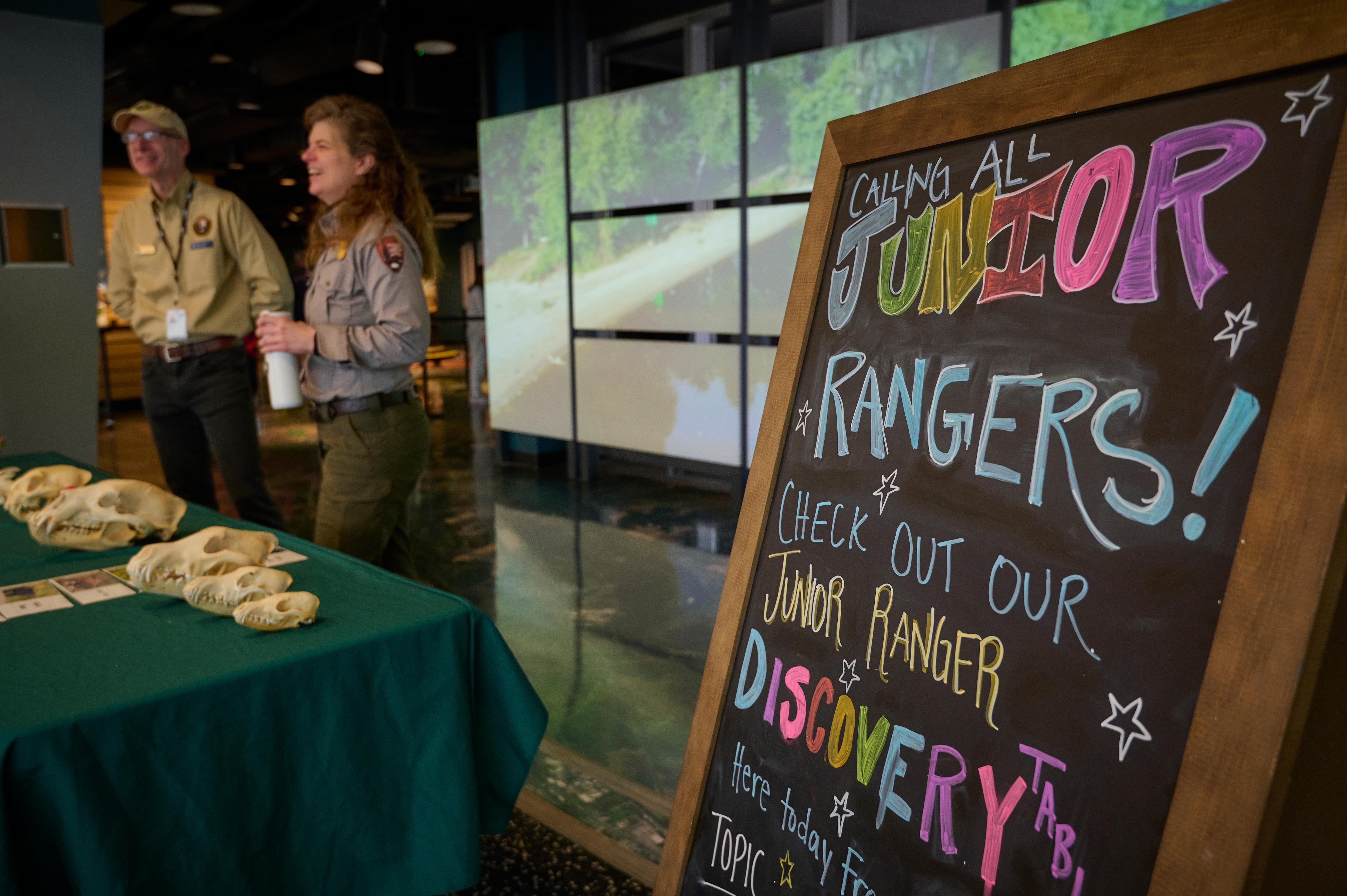 A sign reads "Junior Rangers" with a park ranger and volunteer in the background.
