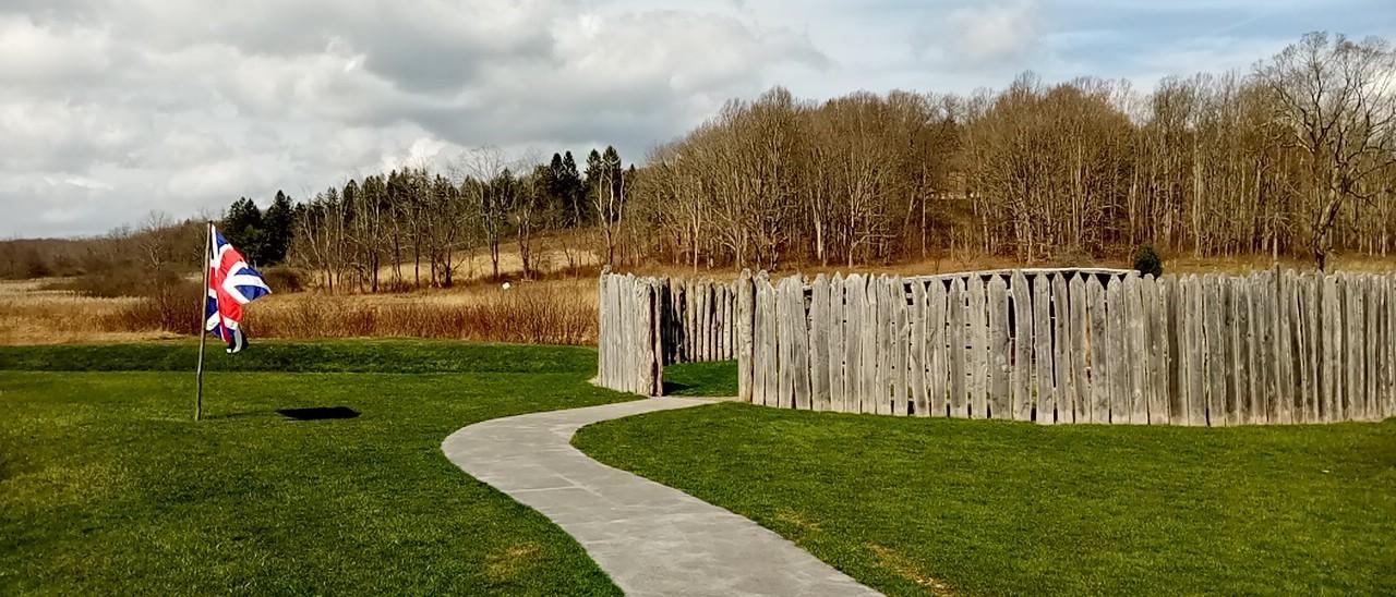 Small circular fort with British flag in a green meadow with bare trees in the background