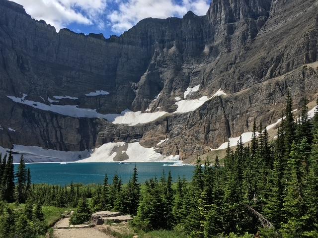 a blue lake is surrounded by forest and a rock cirque with snow on the far side