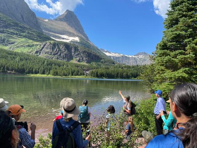 a group of hikers and a ranger look out over a lake with distant mountains and blue sky