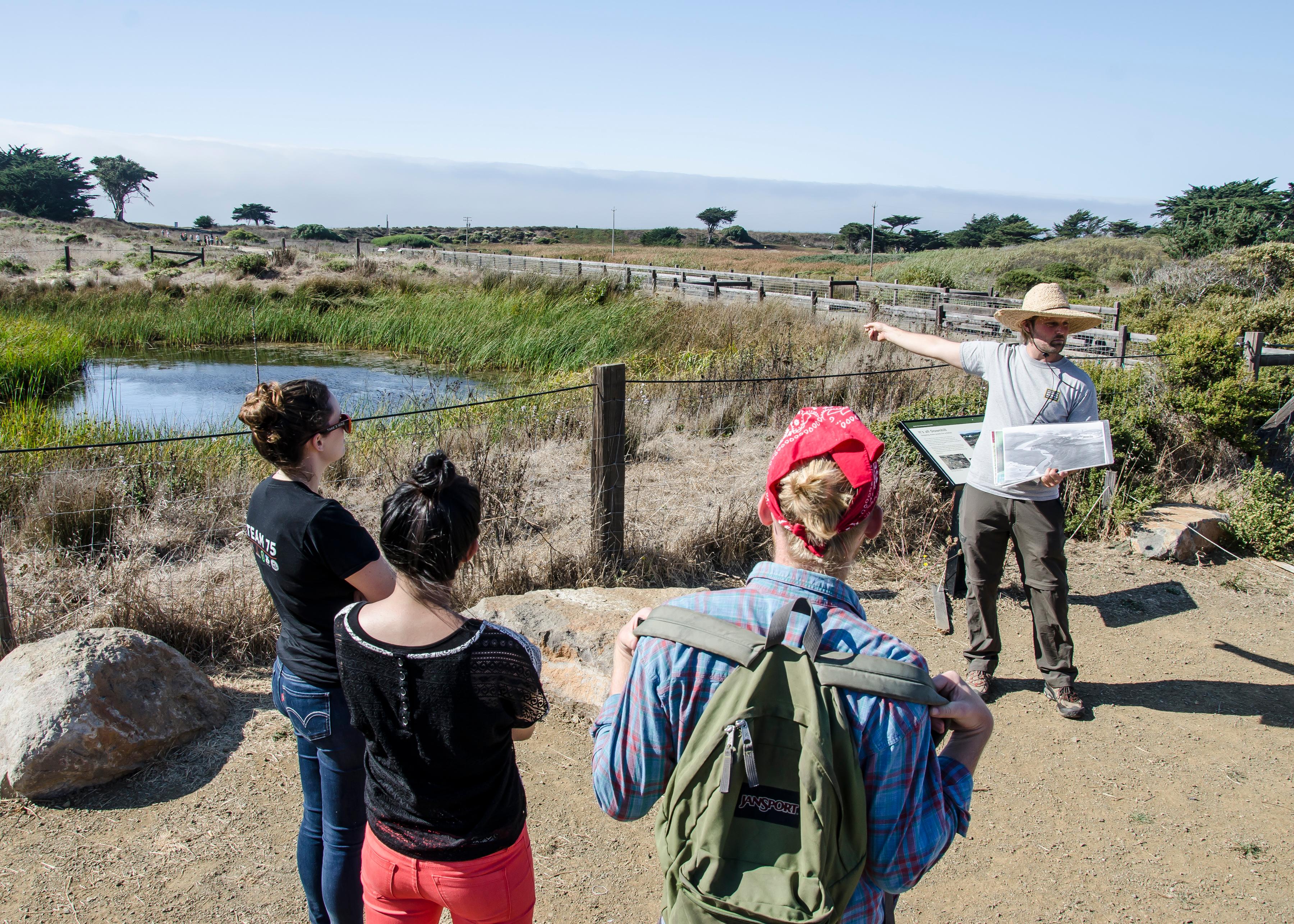 Three visitors look at a man in a broad brimmed hat pointing to a pond while holding an explanatory