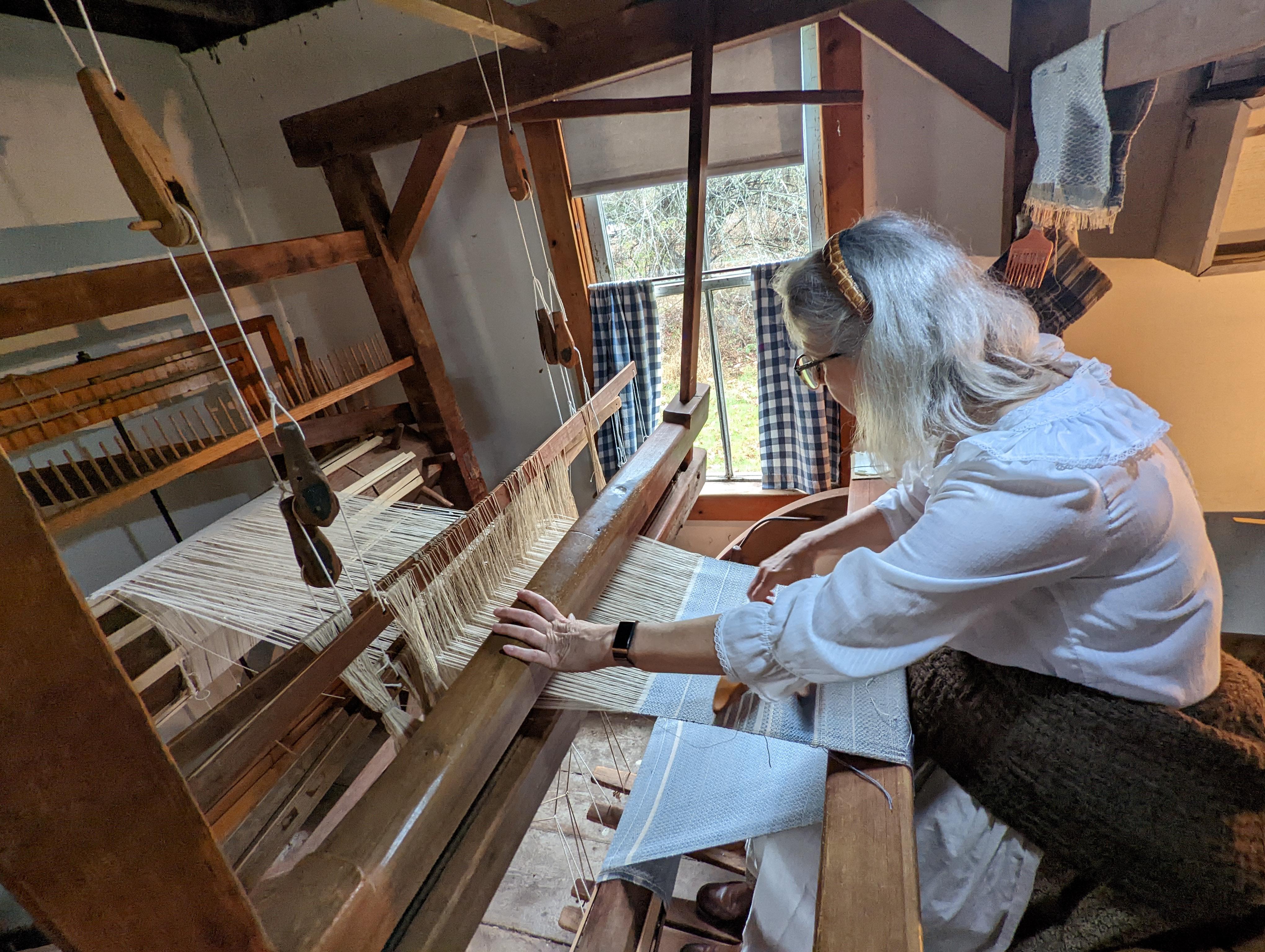A weaver in 19th century clothing at a large loom used for weaving fabric.