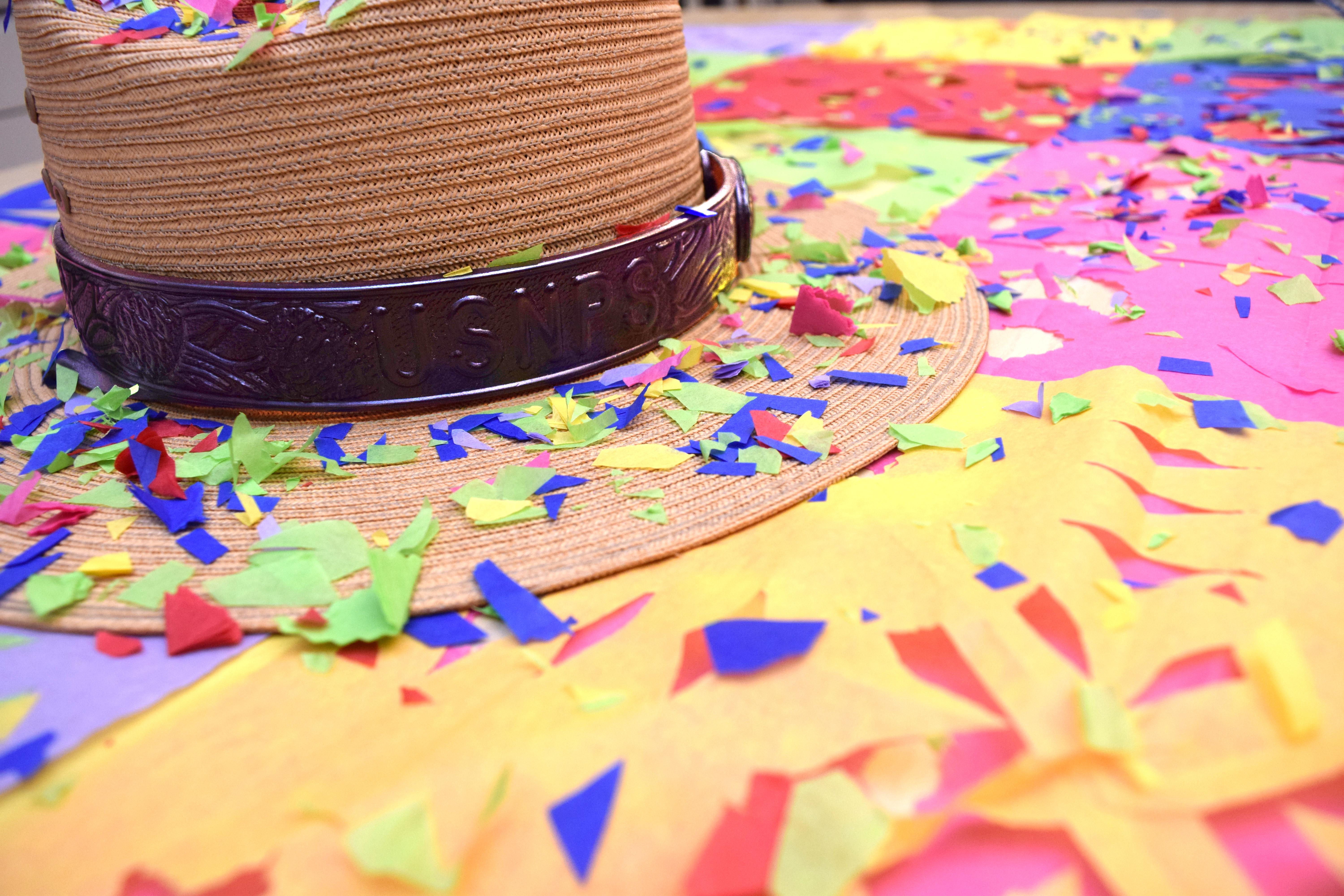 A straw ranger hat with a dark band stamped with &ldquo;USNPS,&rdquo; lying on a colorful surface with confetti.