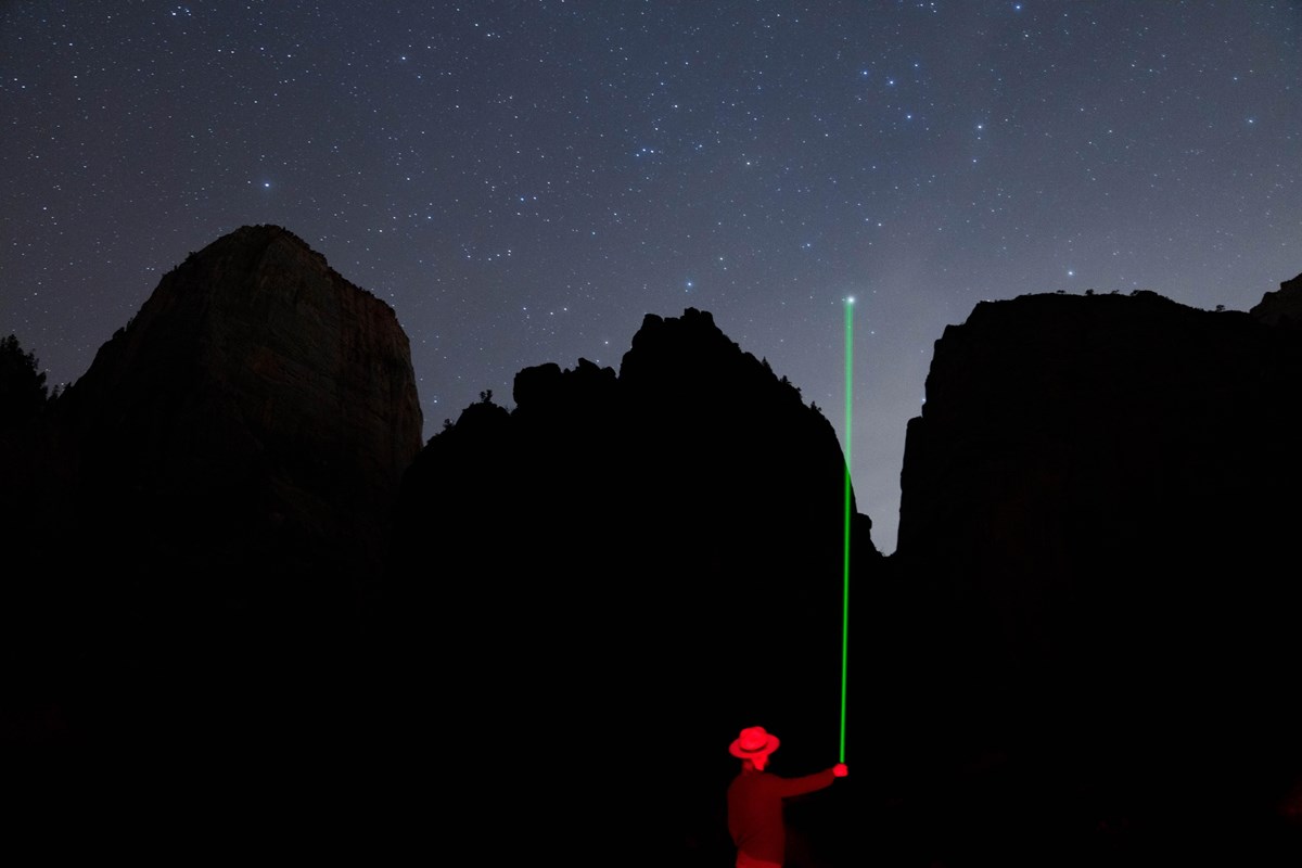 A ranger points a laser pointer at a bright star, with high cliffs silhouetting a starry sky.