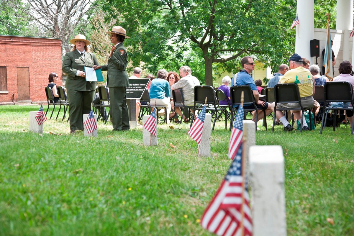 Park Rangers in dress uniform stand beyond a ring of headstones with flags in front of them.