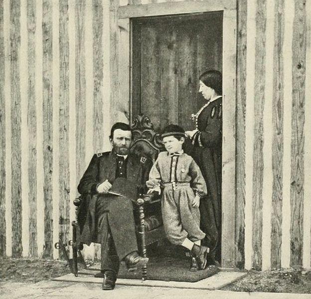 Soldier, woman in a dress, and a young boy in front of a vertical log cabin.