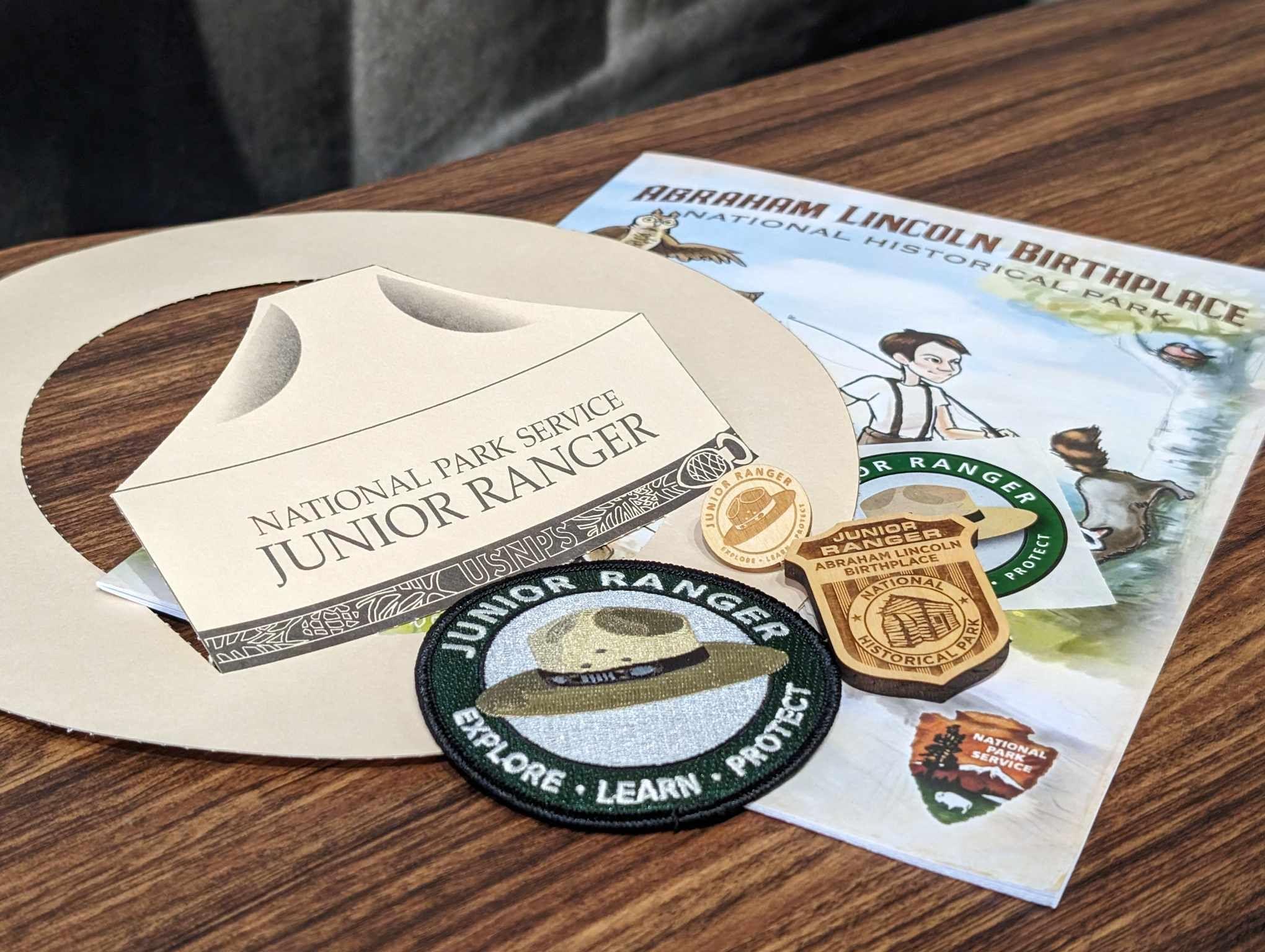 Array of different Junior Ranger swag which includes a book, paper hat, patch, badge, pin & sticker.