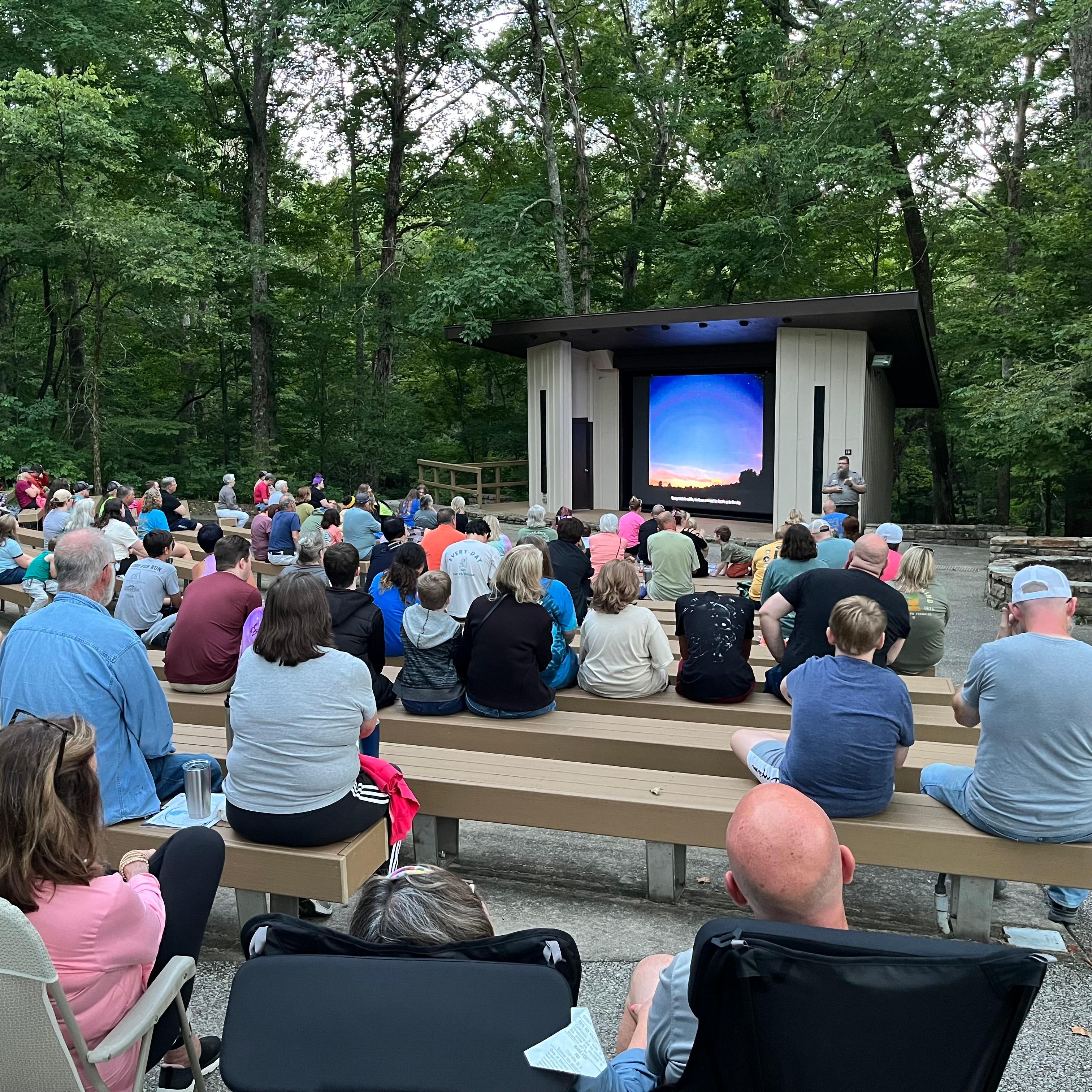 A group of people sit in an outdoor amphitheater with a large screen at the front.