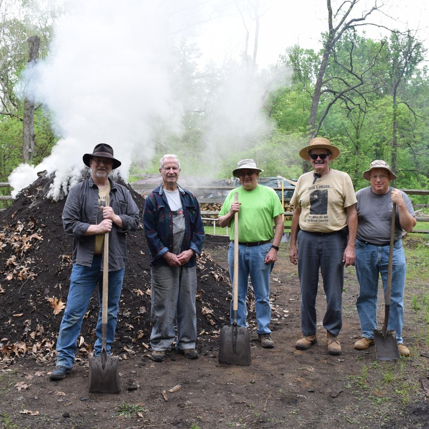 5 men with shovels stand next to burning charcoal pit