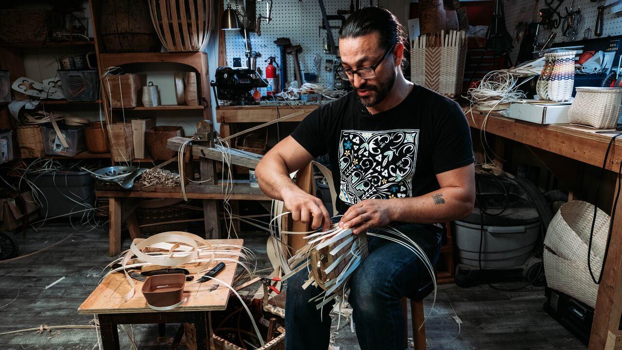 A seated person weaves a basket from strips of brown ash.