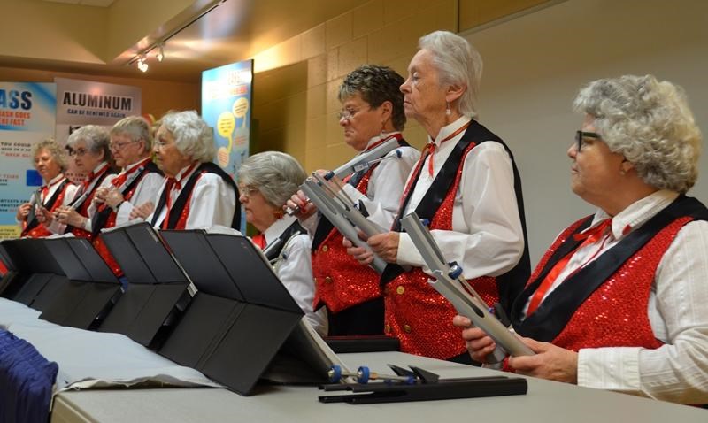 Ten women white shirts with red and black vests and red bow ties playing bells.