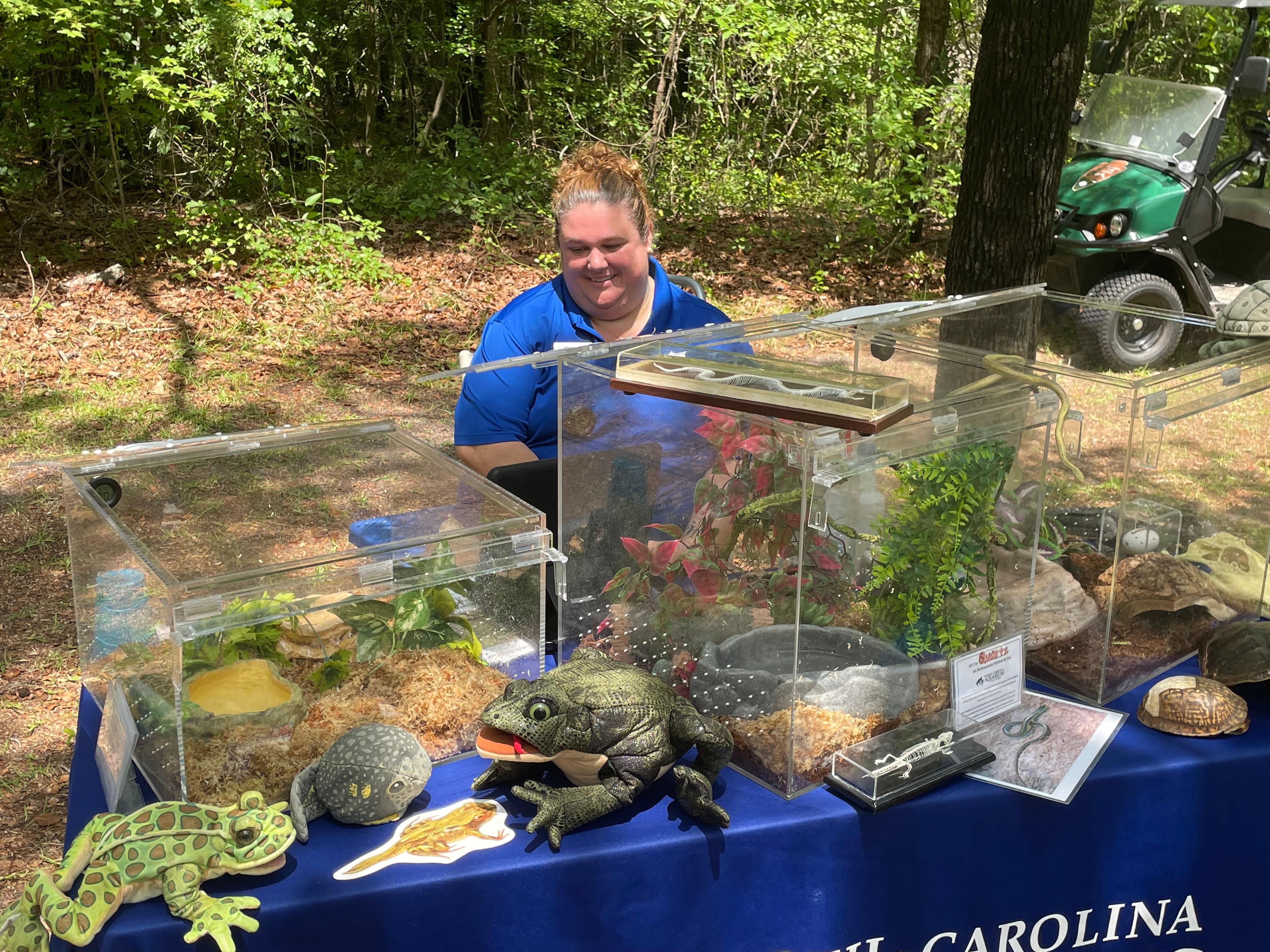 Woman in a blue polo shirt sits at a table with a variety of amphibians and reptiles in glass cages