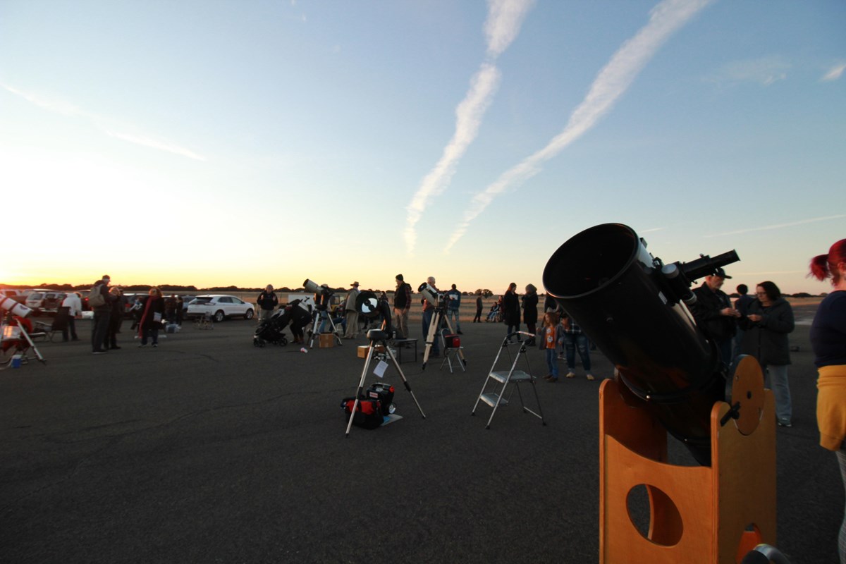 A crowd of people stand around telescopes on a wide, asphalt surface.
