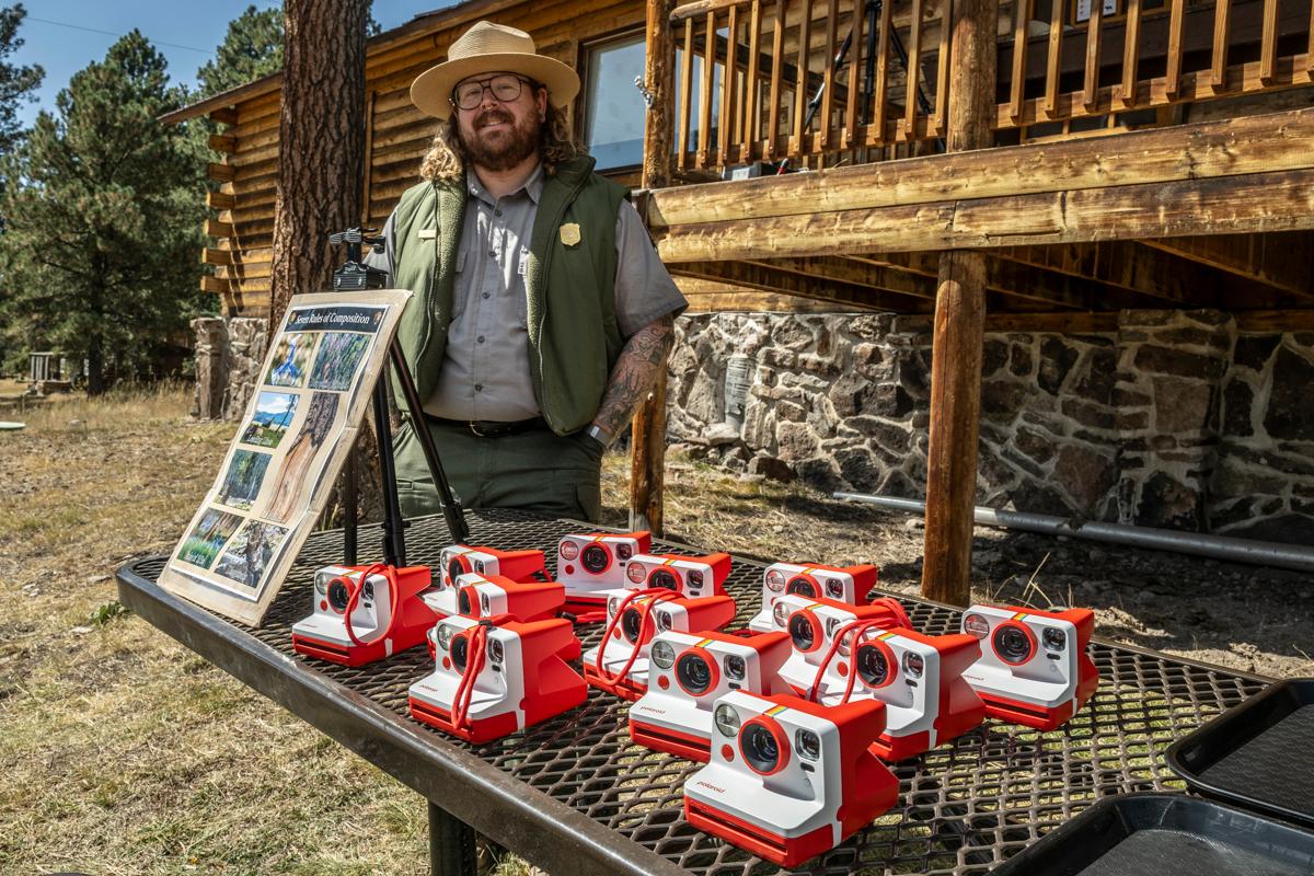 A park ranger poses next to a picnic table filled with red and white instant film cameras.