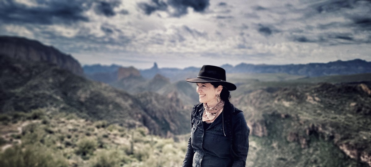 A woman wearing a dark wide-brimmed hat stands in the foreground; behind her is a vast landscape.