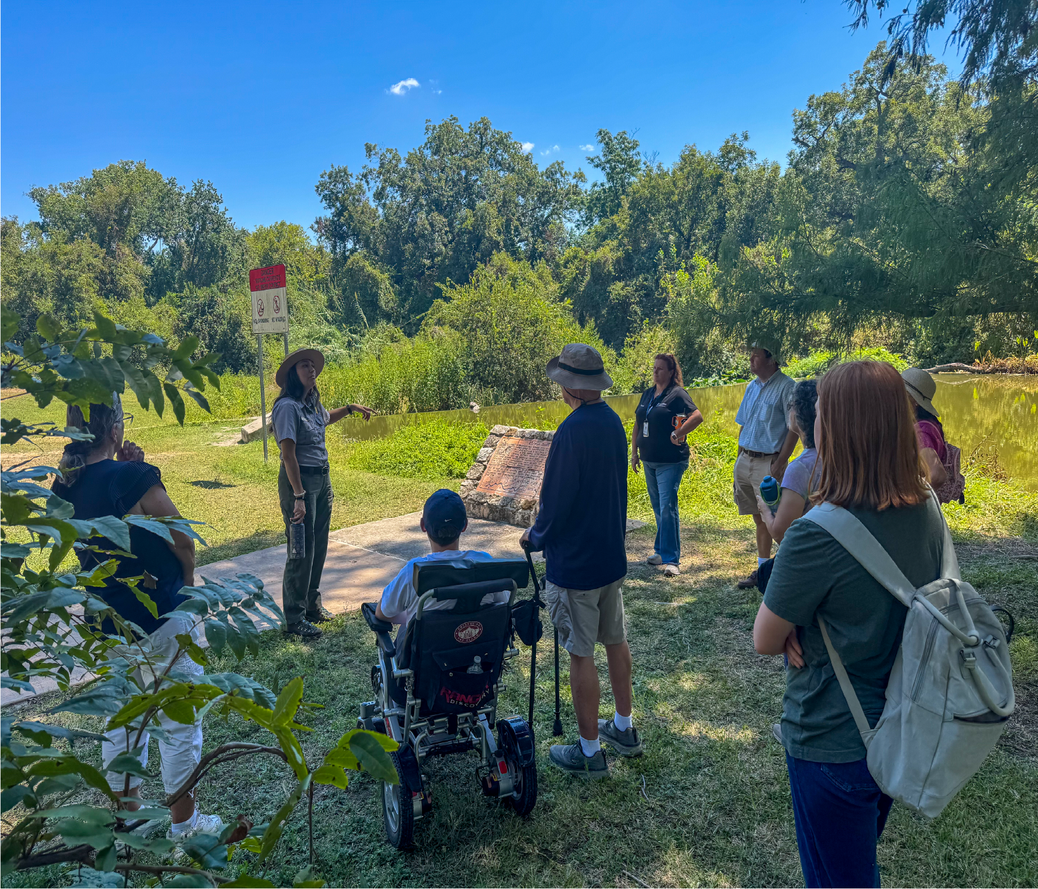 A park ranger stands on a grassy path speaking to a small group of people gathered around outdoors.