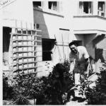 Man in white work suit bends over, holding the paw of a black dog. Both stand in a patio garden
