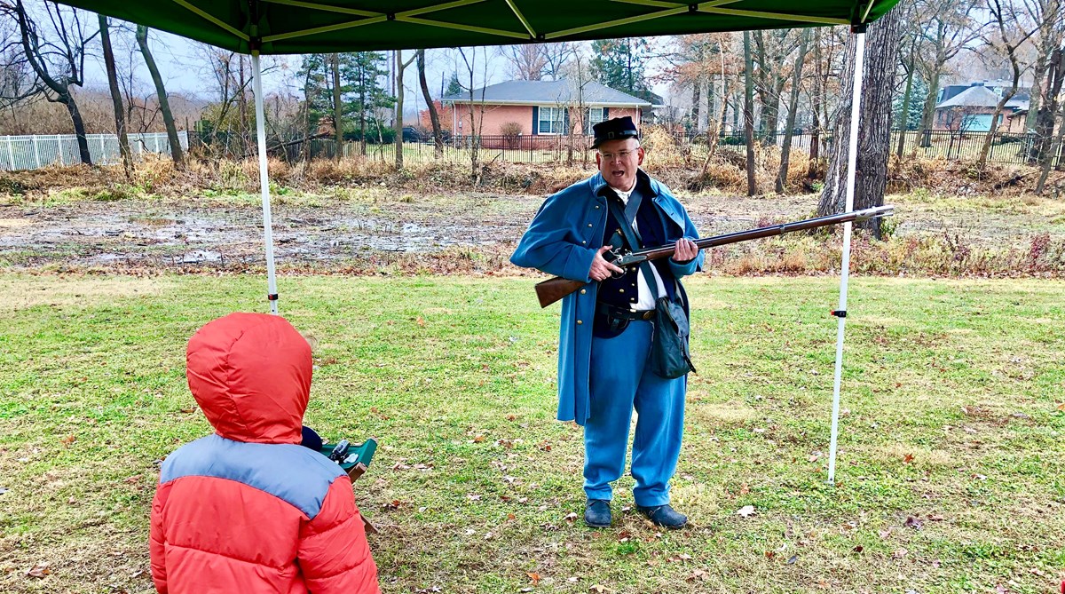 Man in blue Civil War Union uniform holding rifle. Back of young person looking at man.